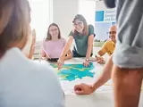 Mehrere Menschen sitzen an einem Tisch. Auf dem Tisch liegt ein großer Plan. Darauf steht ein Windrand. Jemand deutet mit dem Finger auf den Plan.